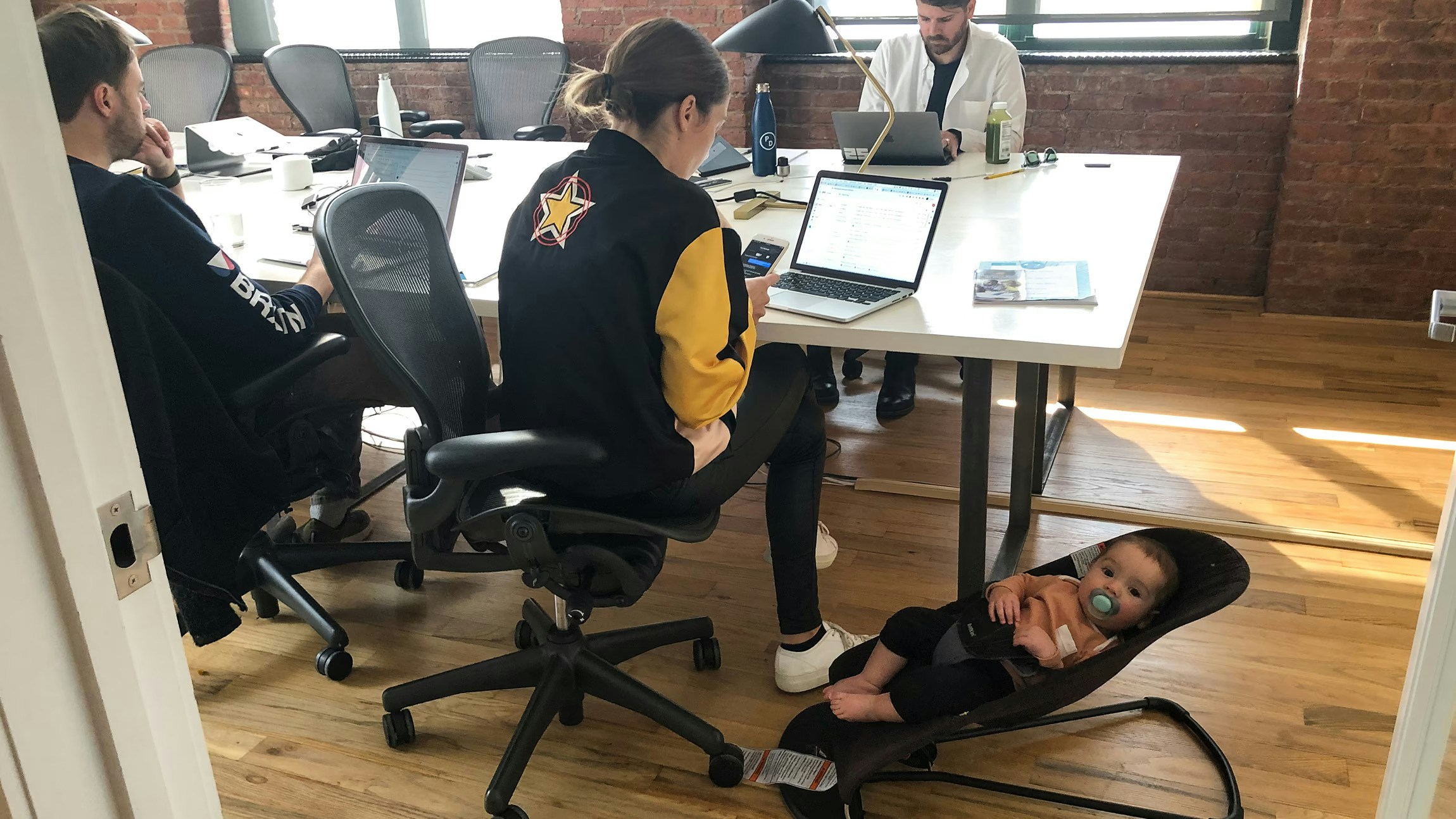Office scene with people working at laptops and a baby in a bouncer chair on the floor