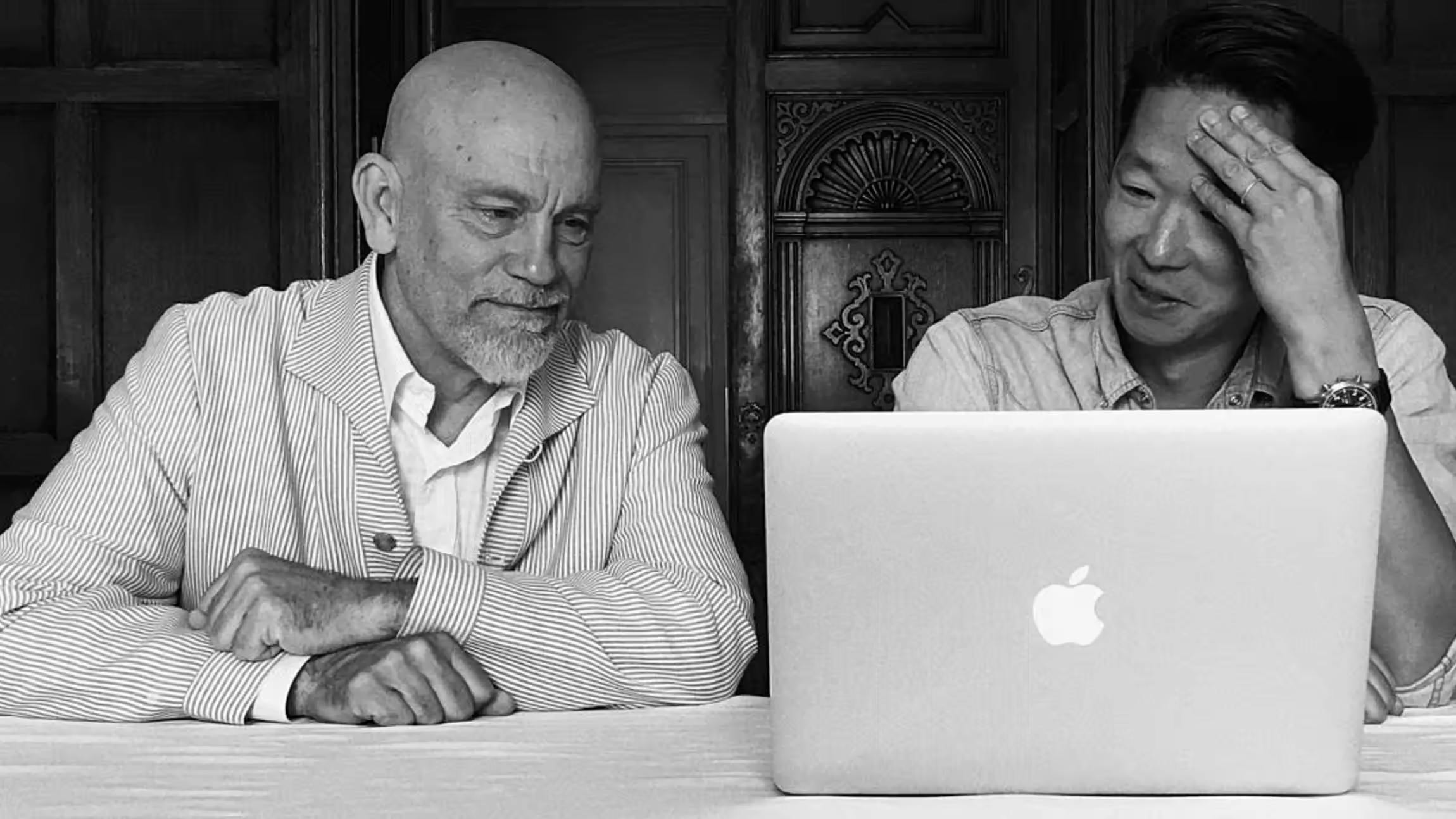 Actor John Malkovich and a colleague sitting at a table, smiling while looking at a laptop screen during a creative meeting