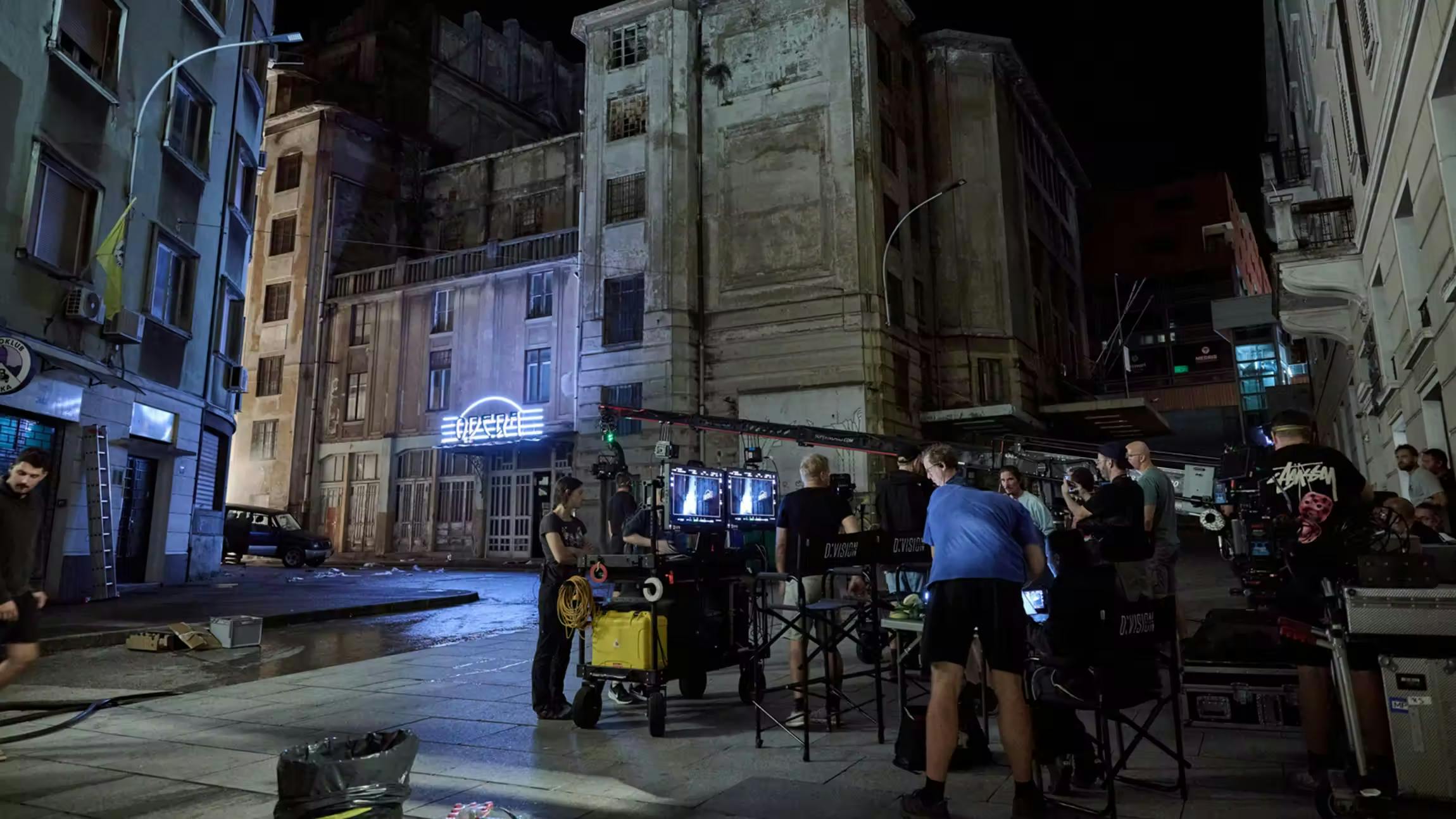 Film crew shooting a night scene on a gritty urban street set with weathered buildings and neon signage