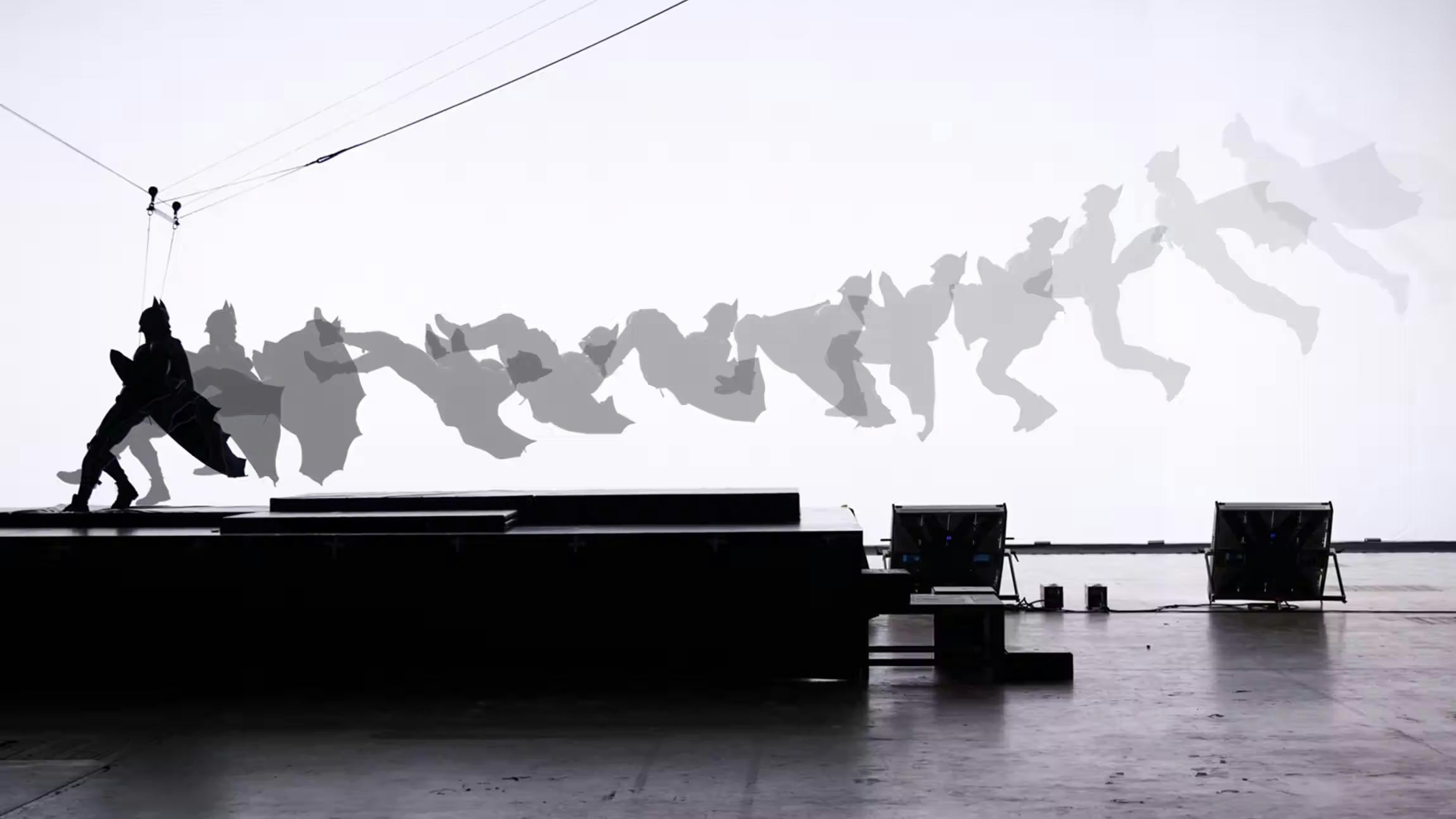 Silhouette of a stunt performer in a Batman costume attached to wires, with multiple shadowed action poses showing a leap in sequence against a white background
