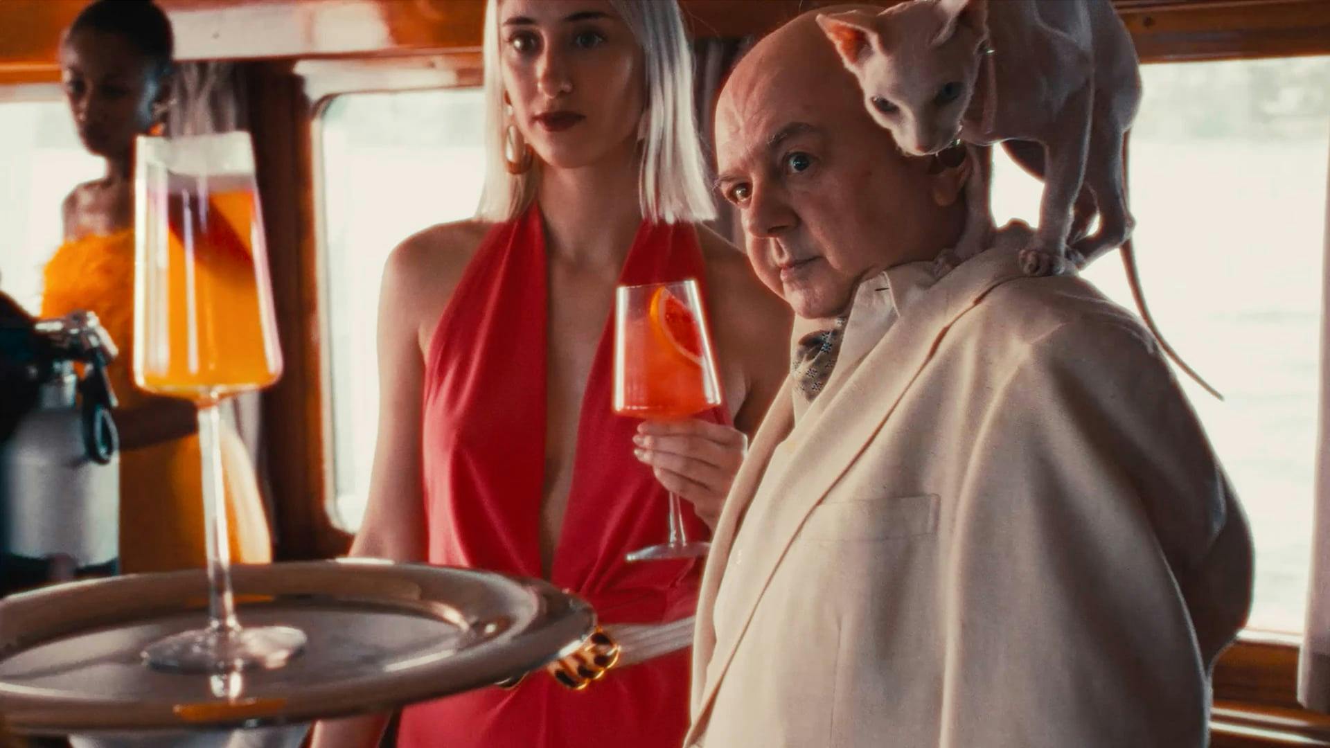 Elegant yacht party scene with man in cream suit, Sphynx cat on shoulder, and woman in red dress holding a citrus cocktail